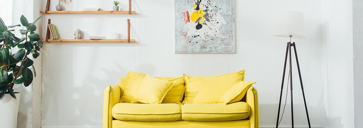 Ein Zimmer mit einem gelben Sofa, einer Lampe, Pflanzen sowie einem Bild und einem Regal an der Wand | Home Staging