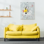 Ein Zimmer mit einem gelben Sofa, einer Lampe, Pflanzen sowie einem Bild und einem Regal an der Wand | Home Staging