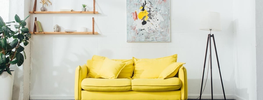 Ein Zimmer mit einem gelben Sofa, einer Lampe, Pflanzen sowie einem Bild und einem Regal an der Wand | Home Staging