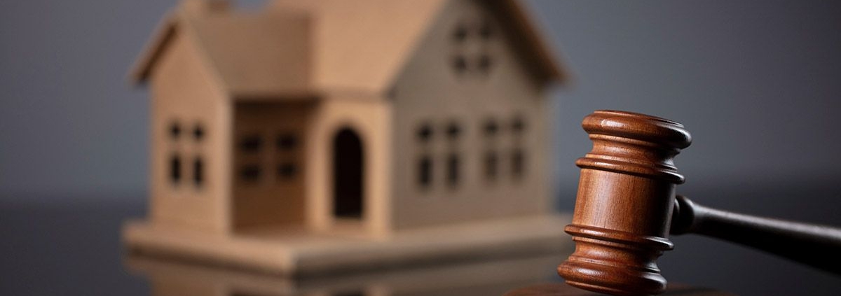 Ein Gavel, oder Richterhammer, aus Holz liegt auf seinem Resonanzblock aus Holz auf einem glänzenden schwarzen Tisch und im Hintergrund steht ein Hausmodell aus Holz | Lebenslanges Wohnrecht