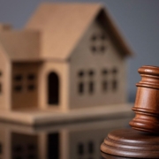 Ein Gavel, oder Richterhammer, aus Holz liegt auf seinem Resonanzblock aus Holz auf einem glänzenden schwarzen Tisch und im Hintergrund steht ein Hausmodell aus Holz | Lebenslanges Wohnrecht