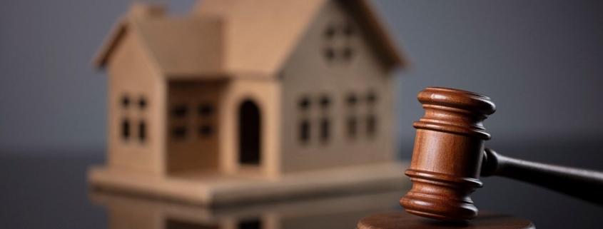 Ein Gavel, oder Richterhammer, aus Holz liegt auf seinem Resonanzblock aus Holz auf einem glänzenden schwarzen Tisch und im Hintergrund steht ein Hausmodell aus Holz | Lebenslanges Wohnrecht