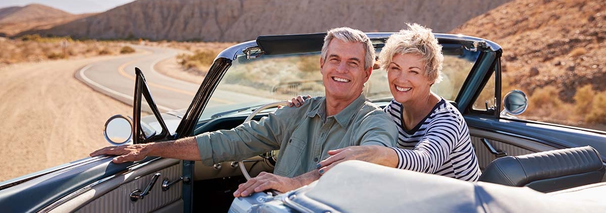 Ein Seniorenpaar sitzt in einem Cabrio auf einer Landstraße in einer weiten Landschaft, die Fahrertür ist auf und beide blicken nach hinten | Immobilienverrentung