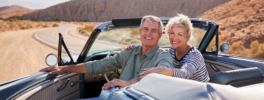 Ein Seniorenpaar sitzt in einem Cabrio auf einer Landstraße in einer weiten Landschaft, die Fahrertür ist auf und beide blicken nach hinten | Immobilienverrentung