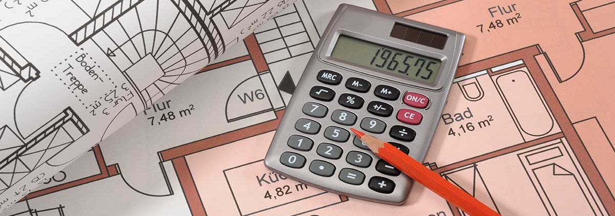 Auf dem Grundrissplan einer Immobilie liegen ein weiterer zusammengerollter Grundrissplan, ein Taschenrechner und ein Stift | Immobilienfinanzierung