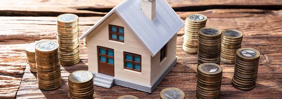 Modell eines Hauses steht auf einem Holztisch und darum stehen kleine Türmchen aus Ein-Euro-Münzen | Immobilienfinanzierung