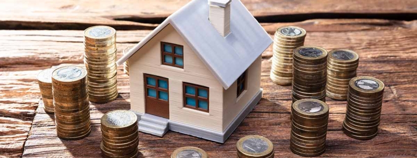 Modell eines Hauses steht auf einem Holztisch und darum stehen kleine Türmchen aus Ein-Euro-Münzen | Immobilienfinanzierung