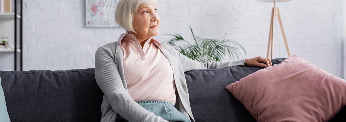 Eine Seniorin sitzt auf ihrem Sofa in ihrem Wohnzimmer, im Hintergrund hängt ein modernes Gemälde, daneben stehen eine Topfpflanze, eine Stehlampe und ein Regal | Wohnen im Alter