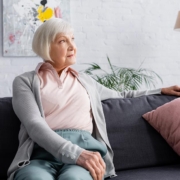 Eine Seniorin sitzt auf ihrem Sofa in ihrem Wohnzimmer, im Hintergrund hängt ein modernes Gemälde, daneben stehen eine Topfpflanze, eine Stehlampe und ein Regal | Wohnen im Alter