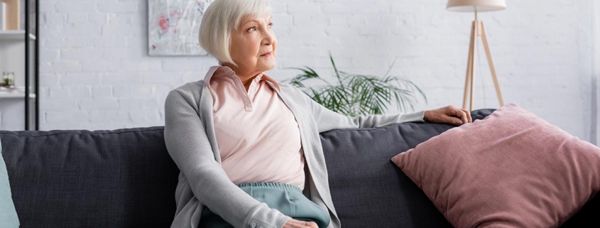Eine Seniorin sitzt auf ihrem Sofa in ihrem Wohnzimmer, im Hintergrund hängt ein modernes Gemälde, daneben stehen eine Topfpflanze, eine Stehlampe und ein Regal | Wohnen im Alter